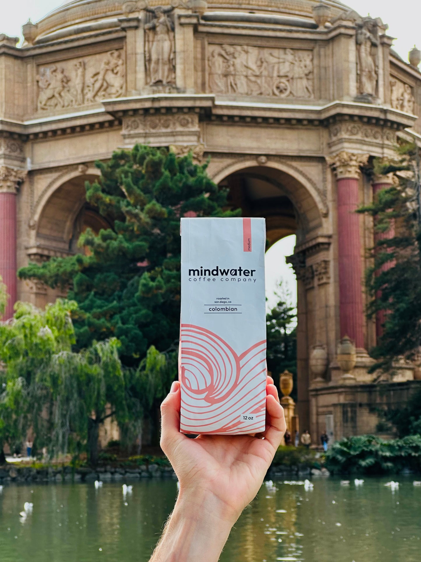 Person holding a 'mindwater' medium roast 12 oz Colombian coffee roasted in San Diego in front of an ornate building in San Francisco with a pond.