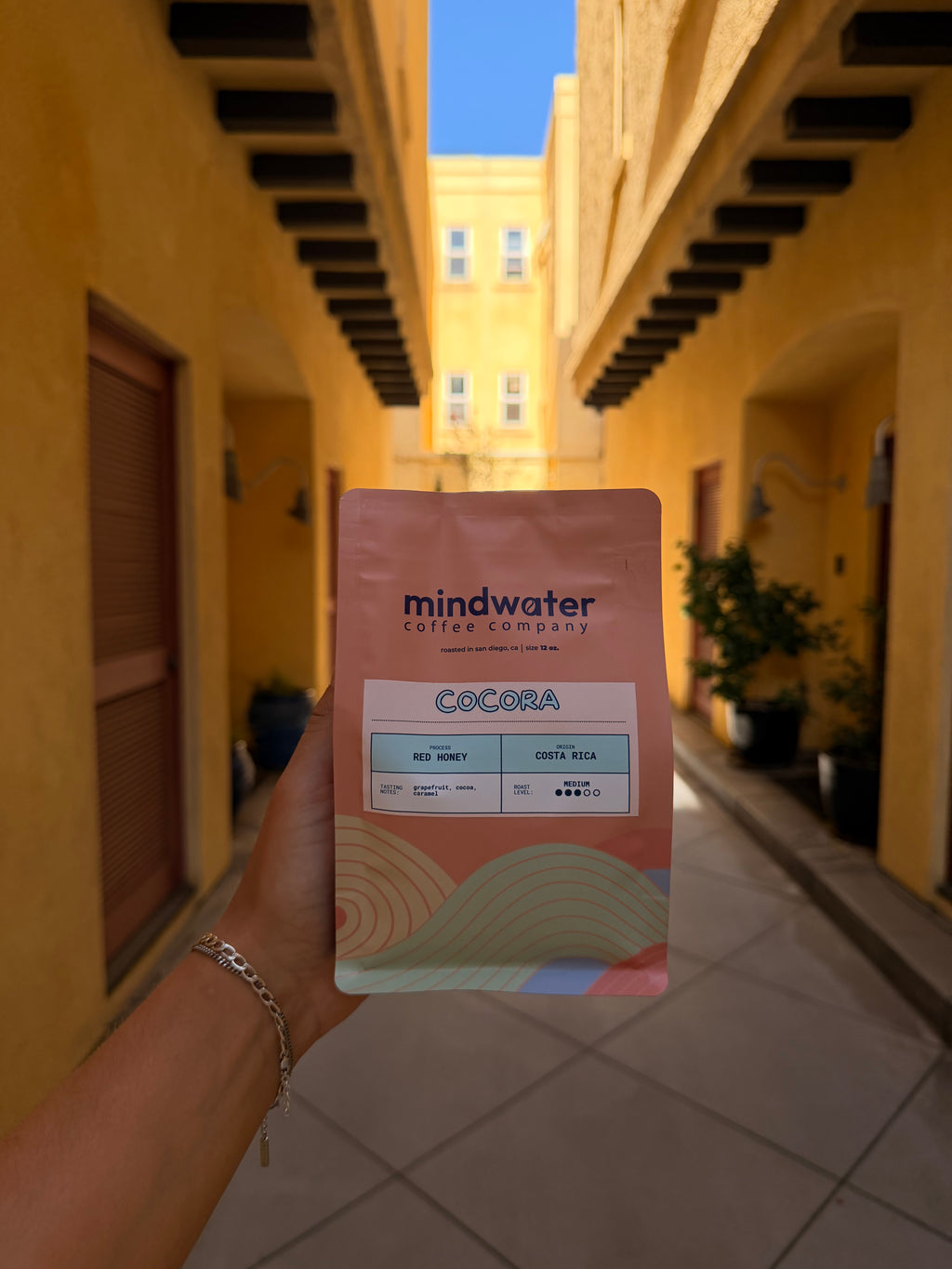 A hand holding a pouch of Mindwater Cocoa coffee in a real-world outdoor setting with a warm yellow building facade in the background.