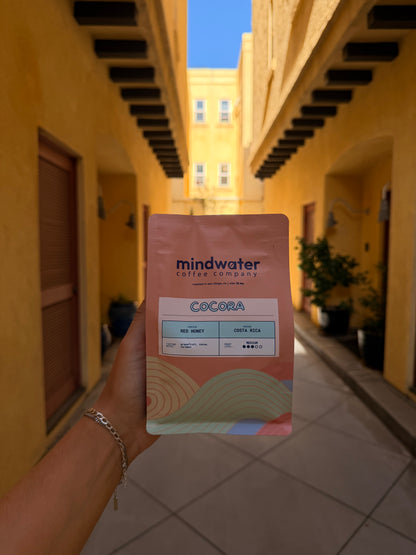 A hand holding a pouch of Mindwater Cocoa coffee in a real-world outdoor setting with a warm yellow building facade in the background.