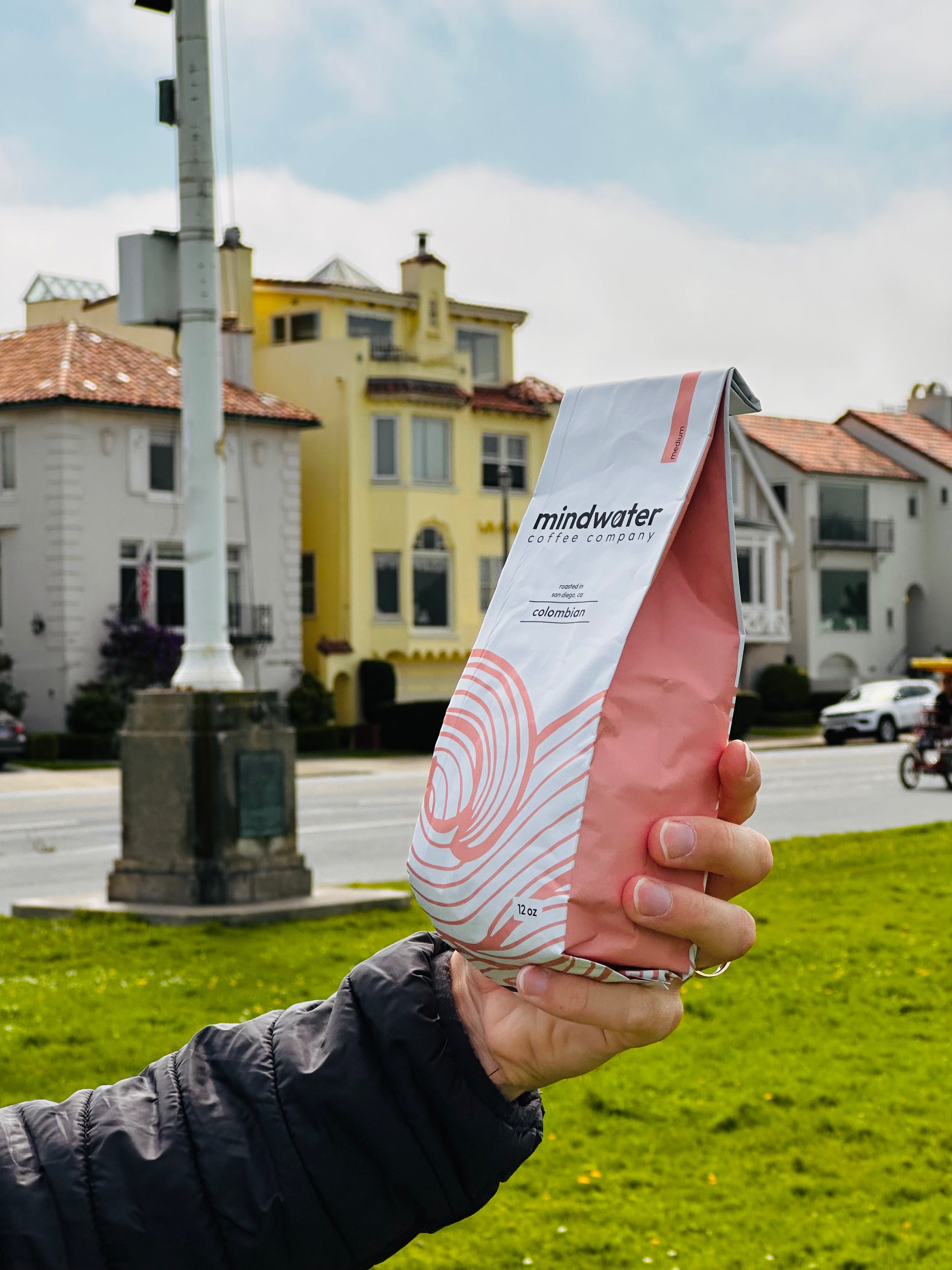 Person holding a pink and white medium roast colombian coffee roasted in San Diego package with 'mindwater' branding in an outdoor setting.