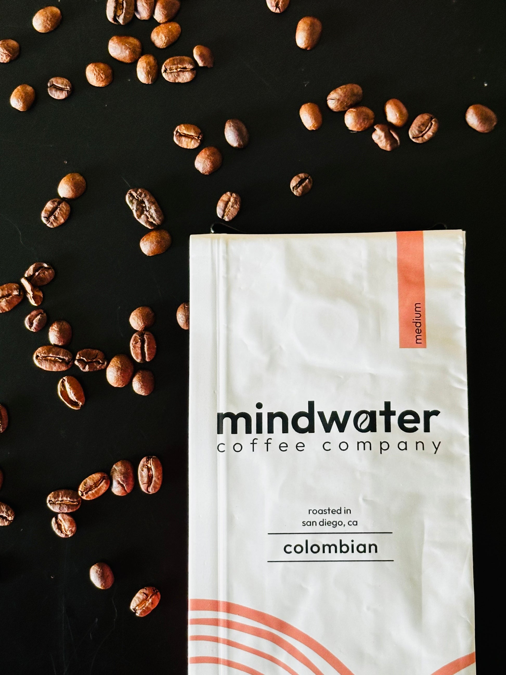 12 oz medium roast colombian mindwater coffee company package roasted in San Diego with scattered coffee beans on a black background