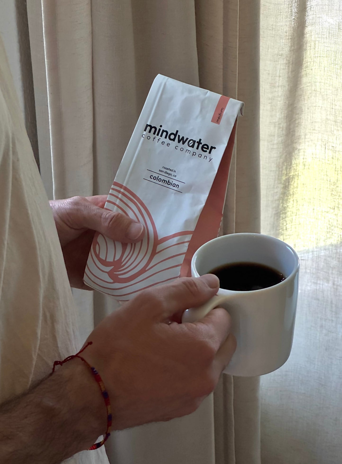 Person holding a white mug with coffee and a 'Mindwater Coffee Company' 12 oz Colombian medium roast roasted in San Diego.