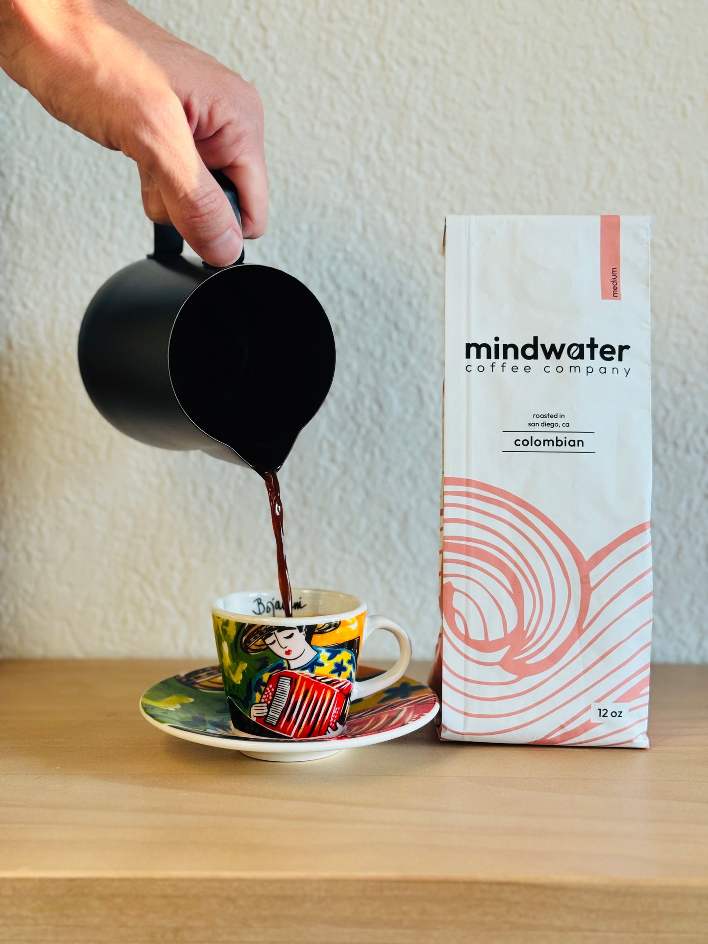 Person pouring coffee into a cup with a Mindwater Coffee Company 12 oz Colombian medium roast coffee on a wooden surface.